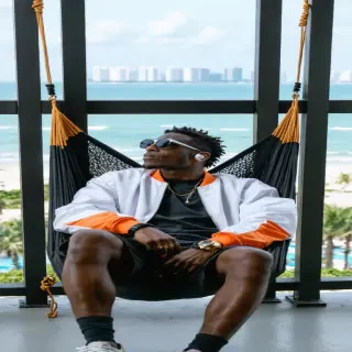 Wide-angle, fashion editorial-style beach shot The monkey lounges on a custom hammock made of black mesh and gold cords, stretched between two modern steel poles on a high-rise balcony overlooking a beach. He wears a designer bomber jacket (white with orange accents), black cargo shorts, high-top sneakers, and reflective mirror shades. Earbuds in, expression calm, he gazes out over the city skyline blending with the sea, exuding a luxury streetwear vibe. His watch flashes subtly in the sunlight — everything about him says cool control.