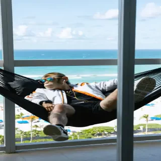 Wide-angle, fashion editorial-style beach shot The monkey lounges on a custom hammock made of black mesh and gold cords, stretched between two modern steel poles on a high-rise balcony overlooking a beach. He wears a designer bomber jacket (white with orange accents), black cargo shorts, high-top sneakers, and reflective mirror shades. Earbuds in, expression calm, he gazes out over the city skyline blending with the sea, exuding a luxury streetwear vibe. His watch flashes subtly in the sunlight — everything about him says cool control.